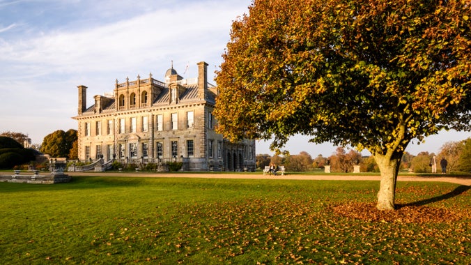 Sun shining on the house at Kingston Lacy, leaves on a tree in the foreground are starting to change colour and drop on the lawn
